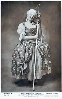 Young white woman in white 18th-century white wig, and costume patterned with flowers; she carries a shepherdess's crook