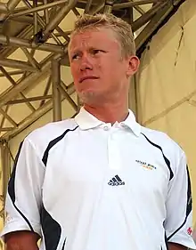 A blond-haired man in his mid-thirties, wearing a white polo shirt bearing an Adidas logo. He is looking to his right.