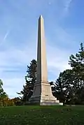 Light colored stone obelisk that stands on a wide base, on which is inscribed family names.