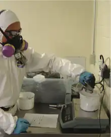 A man wearing a white lab coat reachess over a beaker containing white powder on a balance