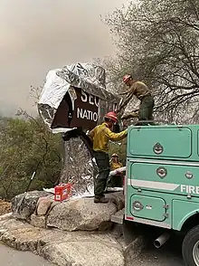 Two firefighters wrap a brown wooden sign with white lettering in sheets of silver foil