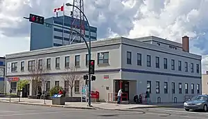 A two-story light gray building seen from across an intersection over which a traffic signal is displaying a red light. A Canadian flag is flying from the flat roof. There are taller buildings behind it.