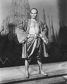 Black and white photo of a man with a shaven head in silky Asian garb; his chest is exposed and his feet are bare; he stands with hands on hips, glowering at the camera