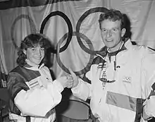 A woman with shoulder length, curly hair and a man with short, curly hair bump fists. They are both in white coats with dark shoulders