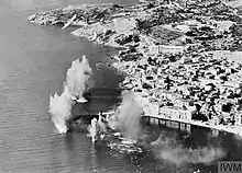 A ship at sea near a town; the ship is surrounded by smoke and spouts of water from explosions