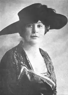 A white woman wearing a large brimmed black hat, and a dark dress with a square neckline