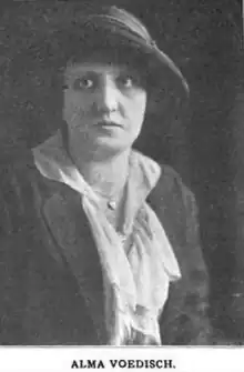 A white woman wearing a slouchy brimmed hat, a dark suit, and a light-colored blouse