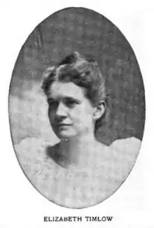 A white woman with hair parted center and dressed to crown; wearing a white dress with a wide ruffled neckline