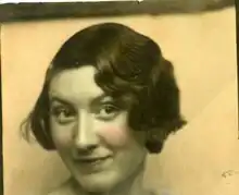 Black and white photo of a woman sitting in a boat with an oar in her hands. She smiles into the camera and has light skin and dark hair.