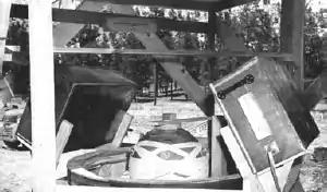 A sphere in a cyclider in the ground, above which is wooden scaffolding and two large boxes. In the background are trees.