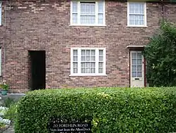 Exterior of a two-story brick building, with a hedge in front of it. Six windows are visible, three on each level, as are two doorways on the lower level.