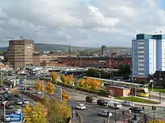 A Metrolink and bus station in front of office buildings. There are hills in the background and a road in the foreground