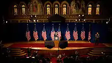 A speaker at a podium in the center of a stage decorated with nine American flags and a red carpet.