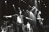 Black and white image of three men holding microphones on a stage. In the background is a drum set, several onlookers, and stage lights shining down from above.