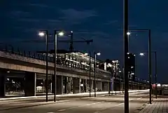 A night view of a viaduct running along the road. In the middle of the image is a glass station.