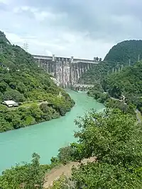 Image of a large structure located between two mountains with power lines, a jungle, a house and a green river