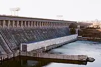 Image of a large concrete structure that looks kind of old and unused with a pier and some structures on it and alongside it