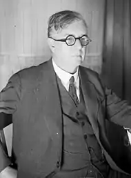 A middle-aged man in a suit, with slightly-unkempt, parted hair and small circular glasses