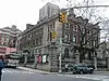 A three story brick and stone building sits on a corner in New York City and is surrounded by a stone and wrought iron fence.