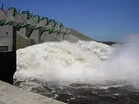 Image of a wall like structure in left, a raging river in the right and power lines located on low hills