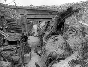 Soldier on guard armed with a rifle wearing a Brodie helmet, three other men are asleep in the bottom of the trench