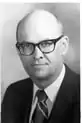 Black-and-white photo of a bald man wearing glasses and a suit with a striped tie