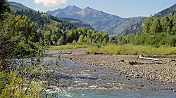 A stream with a rocky shore on the right flows through a hilly, wooded landscape with a high rocky mountain in the distance.