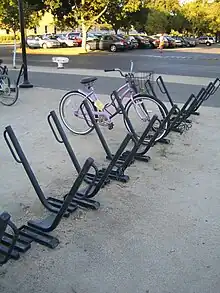 An array of sixteen or so parking spots for bicycles. Each is a square metal bar angled at 45 degrees from the ground with a parallel loop of thinner round tubing at the top. They are set in a row with the bars pointing in alternating directions. A pink ladies' bicycle with a shipping basket is parked in one. Another row of them is mostly out of frame to the left with a mountain bike parked in it. In the background is a car park with trees.