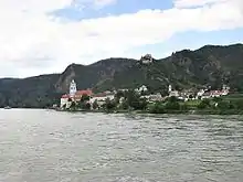 A view of Dürenstein from the river: a small town sits between steep mountains and a wide river. The church tower rises above the several dozen houses. On the mountain above, a ruined castle dominates the sky line.