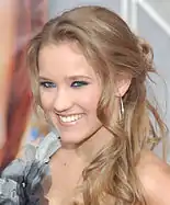 A head shot of a teenage girl with blonde, curly hair, wearing a grey, feathered dress. She is posing at a press event and smiling.