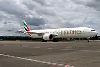 A mostly white Boeing 777, with some red, green and black markings, of Emirates. It is on a runway under a cloudy sky, facing right.