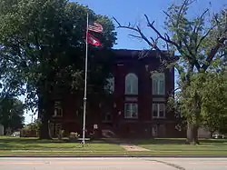 A dark red brick rectangular two and a half-story courthouse hidden behind large trees with a tall flagpole in the front yard