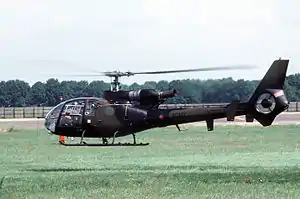 A small camouflaged helicopter hovers, just off the ground, over a field.