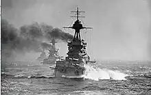 A large gray battleship steams in choppy seas; thick black smoke billows from its funnels. Two battleships are directly behind