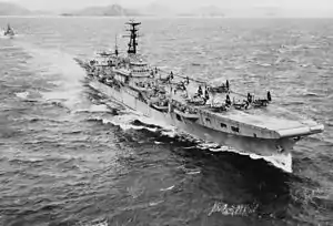 A small aircraft carrier travelling at speed through choppy water. Several propeller aircraft with folded wings are sitting on the flight deck. A small warship is following in the carrier's wake, and a rocky, hilly shoreline can be seen across the horizon