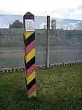 A square wooden post with diagonal black, red and yellow stripes and a metal sign saying "DEUTSCHE DEMOKRATISCHE REPUBLIK" stands in front of a steel mesh fence, through which a guard tower is visible.