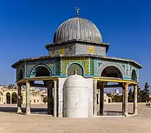 A gray metal-domed octagonal structure decorated with tiles of different colors and geometric designs, supported by dark stone columns with beige-colored capitals