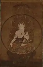 Frontal view of a deity seated on a pedestal in lotus position embellished with ornaments. The right arm is hanging down supported by the legs with the palm of the right hand facing forward. The left hand is placed in front of the body.