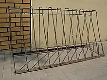 A rusty metal A-frame with horizontal bars at the top and bottom joined by eleven vertical bars. It stands free on concrete in front of a join between a brick wall and a plaster wall.