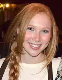 An eighteen year old redheaded caucasoid female is wearing a beige shirt with her ponytail draped over he right shoulder; she is smiling into the camera.