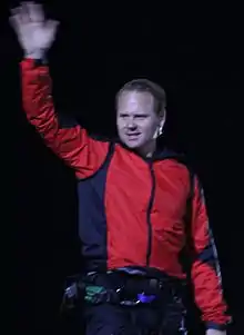 Man in a red jacket waves with his right hand raised high in the air