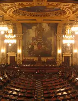 A view of a large, ornately-decorated room with several rows of curved desks, arranged in a semicircle. A large mural is visible on the wall at the far end of the room.