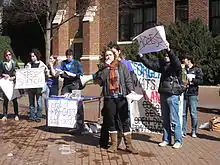 A college-aged female in jacket and scarf holds the microphone attachment of a bullhorn while other students hold protest signs behind her. Two with large red X's over the words read "Free Speech" and "Access."