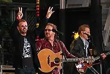 Starr is on stage with two guitarists and two microphones. He is wearing sunglasses and a black and silver t-shirt, and both of his arms are raised. His right arm forms a V-shaped peace symbol.