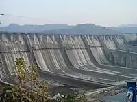 Image of a wall like structure located between mountains with curves at the bottom of it, some kind of plant and a rusty rail bar