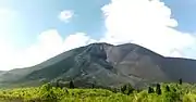 A photograph depicting a blue sky at the top, a grey mountain range in the middle, white clouds in front of the mountain range, and a rocky terrain at the bottom.