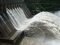 Aerial image of a river flowing from a concrete structure in the shape of an waterfall, with some forest in the background