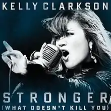 A blue-tint-in-a-black-and-white image of a woman singing. Her right hand is holding a vintage carbon microphone in front of her mouth. The microphone's wire is resting on her left hand between her thumb and her index finger. Below her, the words "Stronger" and "(What Doesn't Kill You)" are written in white upper-case letters.