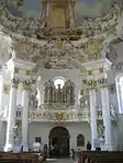 Ornate church interior, looking toward the entrance. The interior is white, the doors flanked by two pairs of columns which stretch to the richly painted ceiling. Above the entrance is the church's pipe organ.