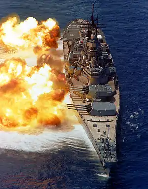 A large ship partially obscured by fire emanating from gun barrels pointed to the left.